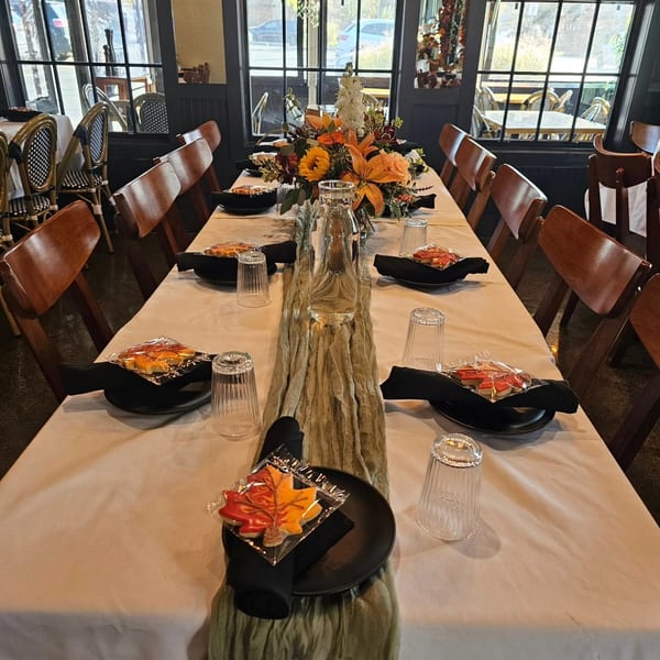 Long dining table set with autumn flowers, black plates, leaf cookies, and wooden chairs in a restaurant.