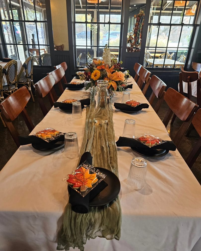 Long dining table set with autumn flowers, black plates, leaf cookies, and wooden chairs in a restaurant.