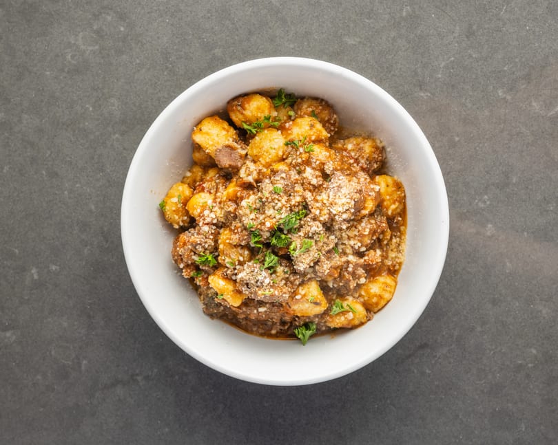 A white bowl filled with gnocchi, meat sauce, grated cheese, and fresh parsley on a dark surface.