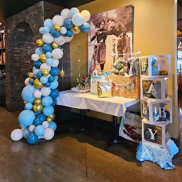 Blue, white, and gold balloon arch with a gift table and "BABY" blocks in a restaurant.