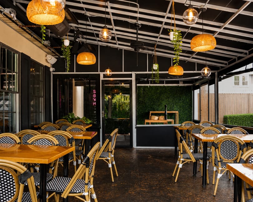 Spacious outdoor restaurant patio with wooden tables, woven chairs, and hanging wicker lights.