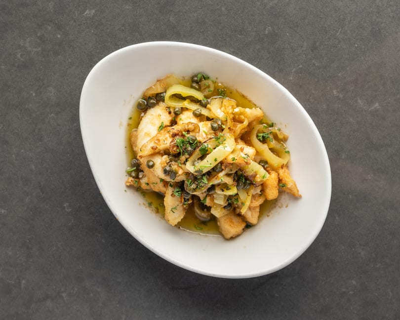 Fried calamari with capers, onions, and parsley in a white bowl on a dark surface.