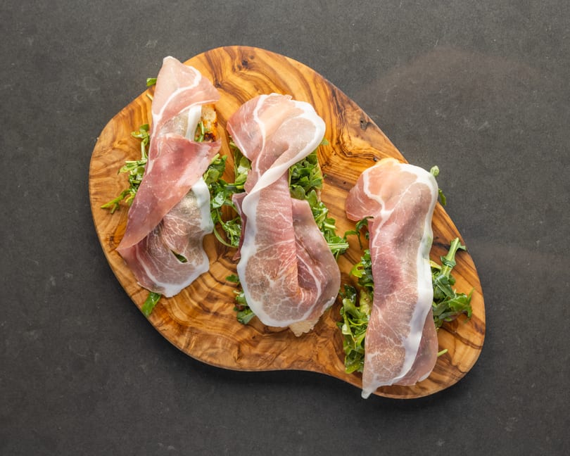 Three prosciutto and arugula bruschetta on a wooden serving board against a dark background.