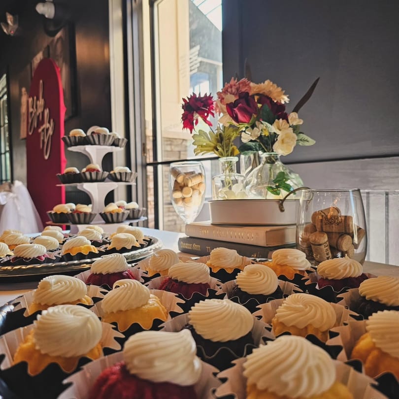 A table filled with frosted mini cakes, decorative flowers, books, and wine corks.
