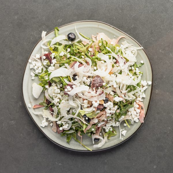 Overhead view of a fresh salad with arugula, feta, olives, and shaved fennel on a gray plate.