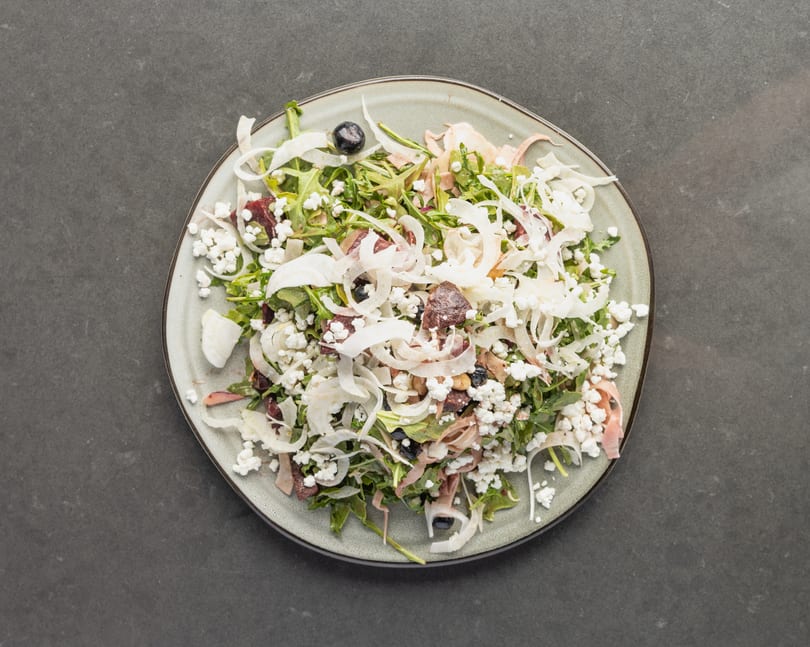 Overhead view of a fresh salad with arugula, feta, olives, and shaved fennel on a gray plate.