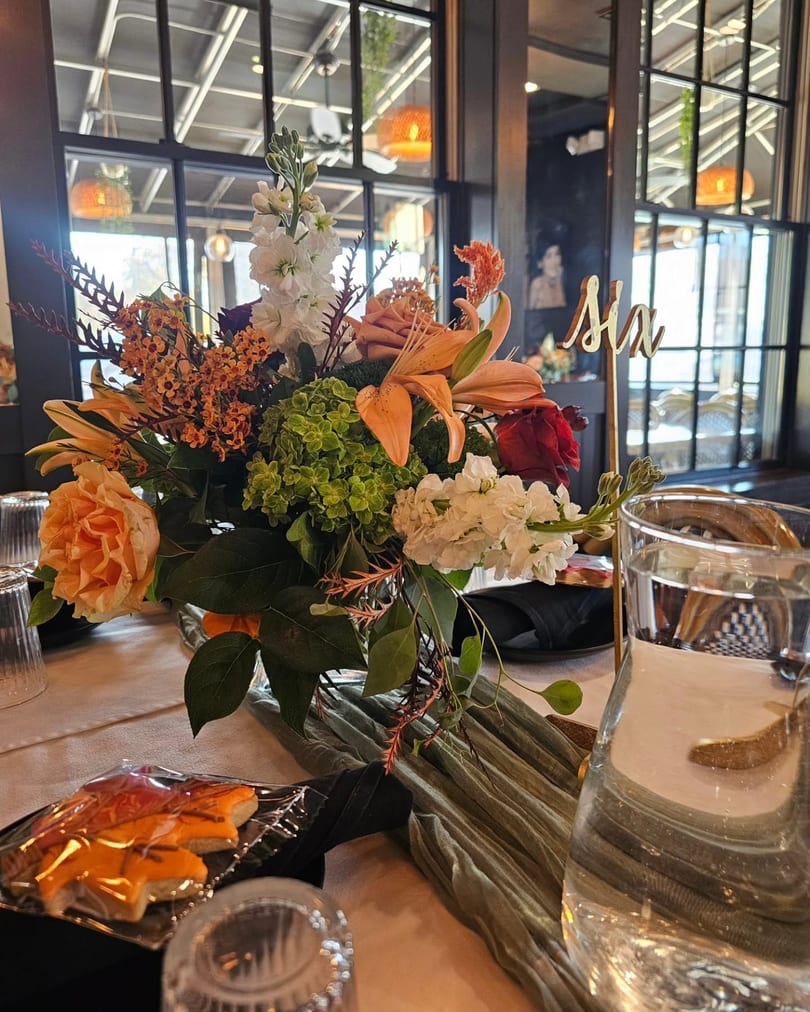 Vibrant floral centerpiece with orange, white, and green flowers on a restaurant dining table with a 'Six' sign.