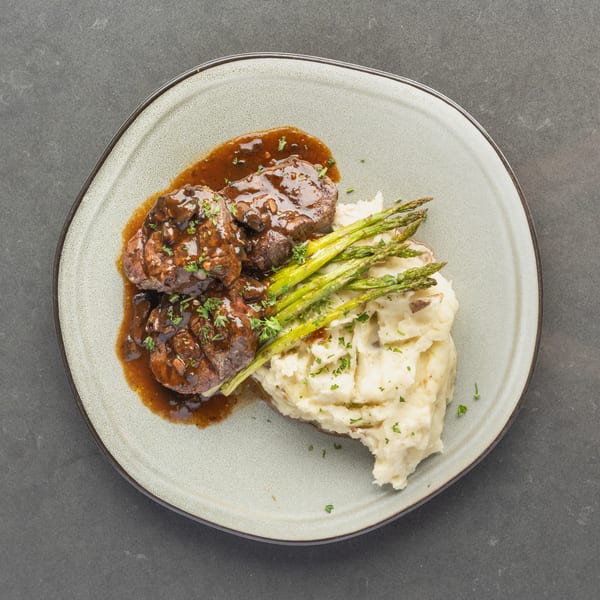 Three pieces of steak with gravy, mashed potatoes, and asparagus on a light gray plate.