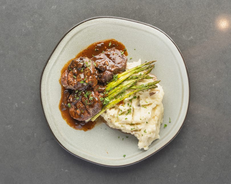 Three pieces of steak with gravy, mashed potatoes, and asparagus on a light gray plate.