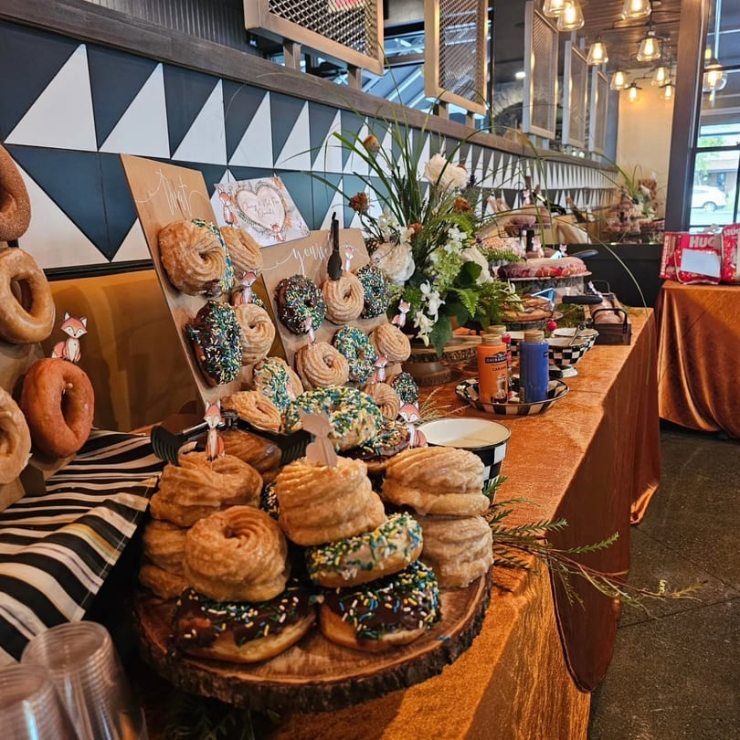 Restaurant dessert table featuring an array of glazed, sprinkled, and cruller donuts with floral decorations.