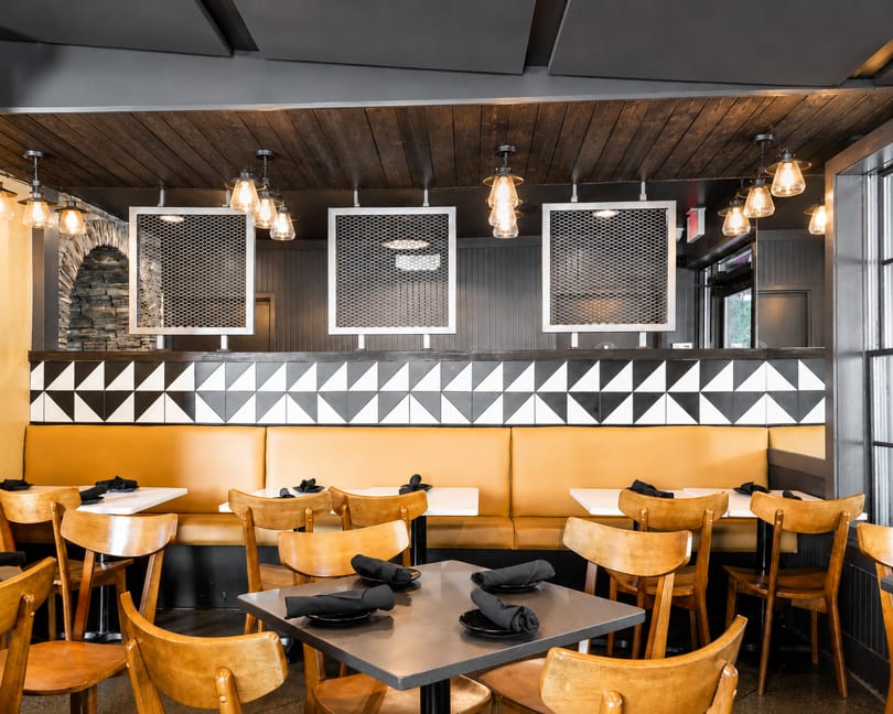 Modern restaurant interior with yellow booths, wooden chairs, geometric wall tiles, and industrial lighting.