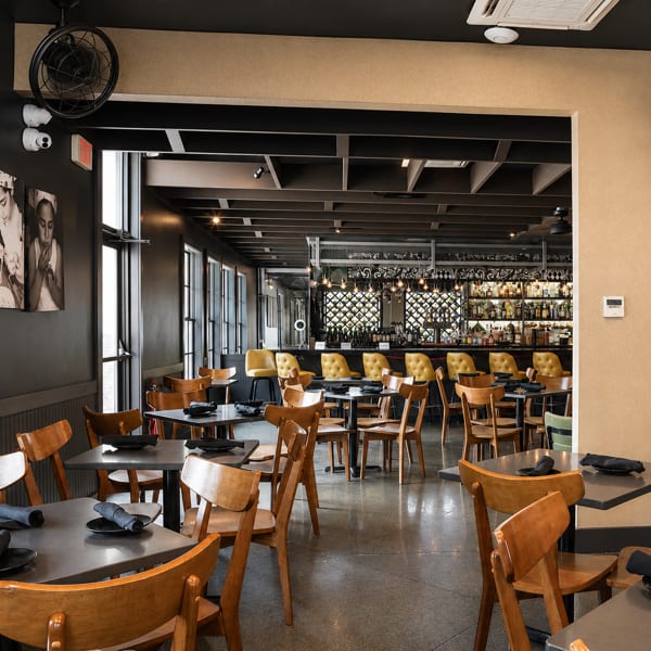 Modern restaurant interior with dining tables, wooden chairs, and a well-stocked bar with yellow stools.