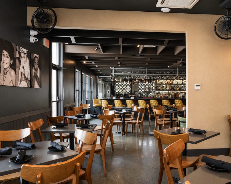 Modern restaurant interior with dining tables, wooden chairs, and a well-stocked bar with yellow stools.