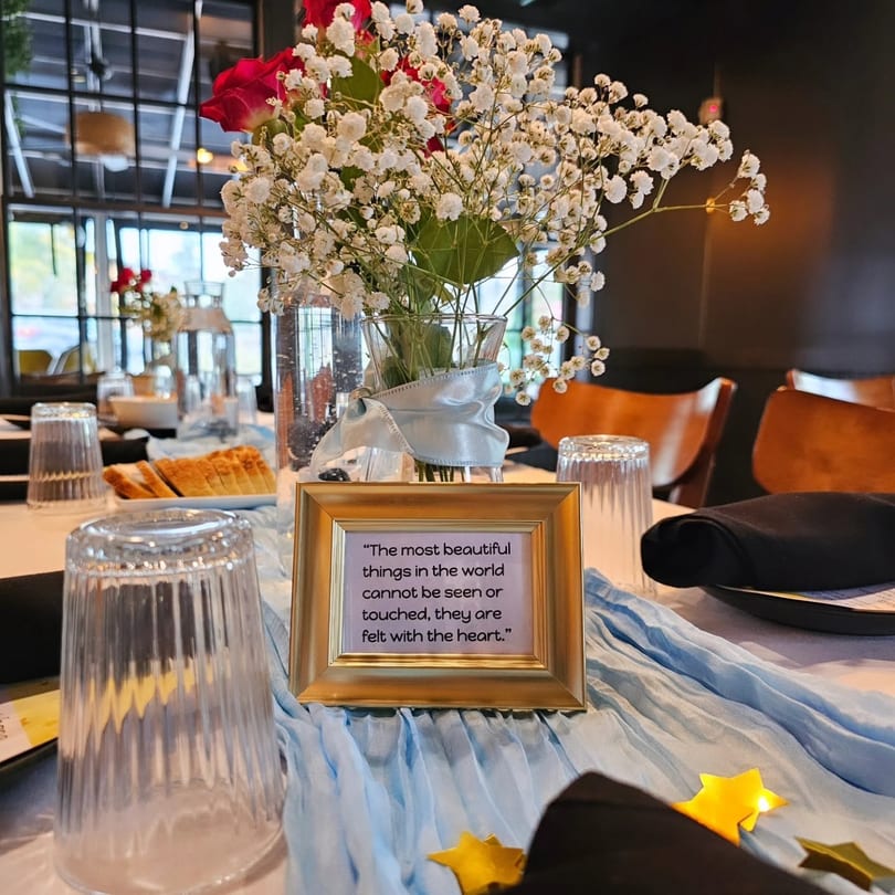 Restaurant table set with a floral centerpiece, blue runner, and a framed inspirational quote.