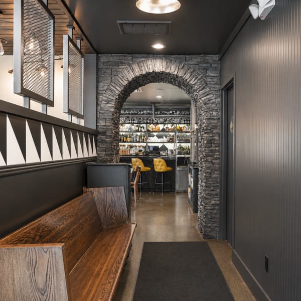 A restaurant hallway with a wooden bench, black and white wall pattern, and a stone archway to a bar.
