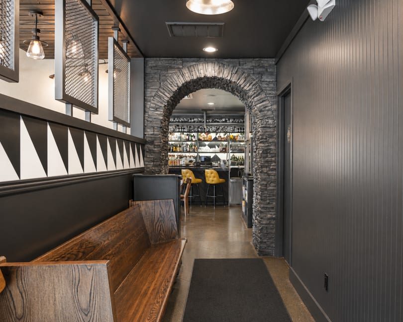 A restaurant hallway with a wooden bench, black and white wall pattern, and a stone archway to a bar.