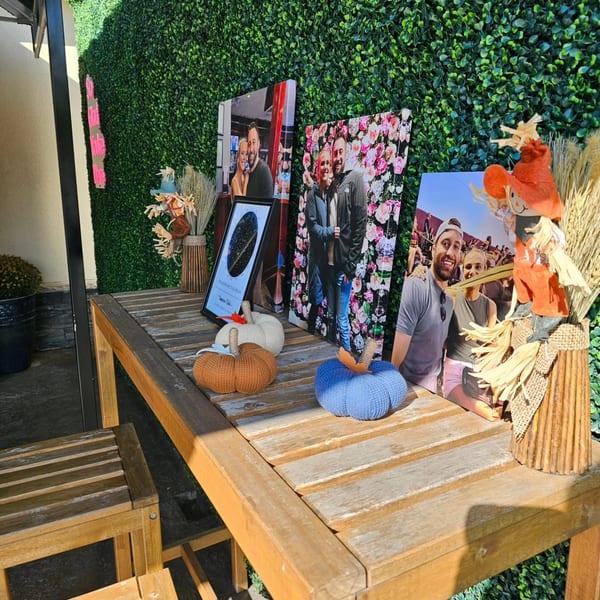 Outdoor wooden table decorated with framed couple photos, knitted pumpkins, and a scarecrow by a hedge wall.