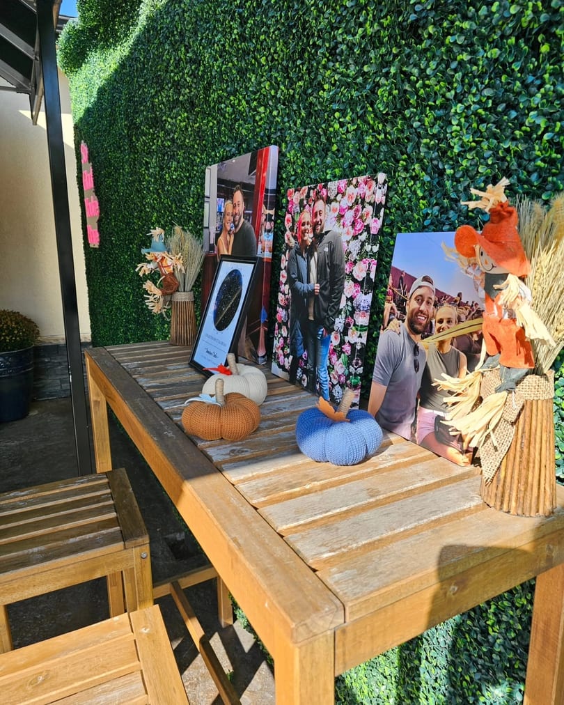 Outdoor wooden table decorated with framed couple photos, knitted pumpkins, and a scarecrow by a hedge wall.