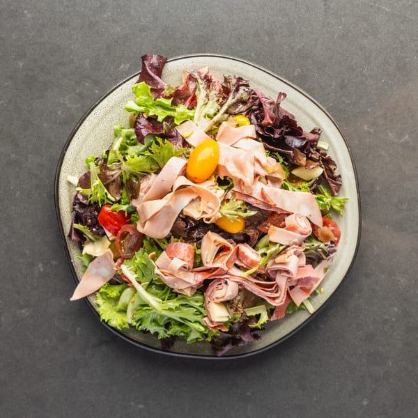 Overhead view of a salad with mixed greens, deli meats, cherry tomatoes, and cheese on a plate.