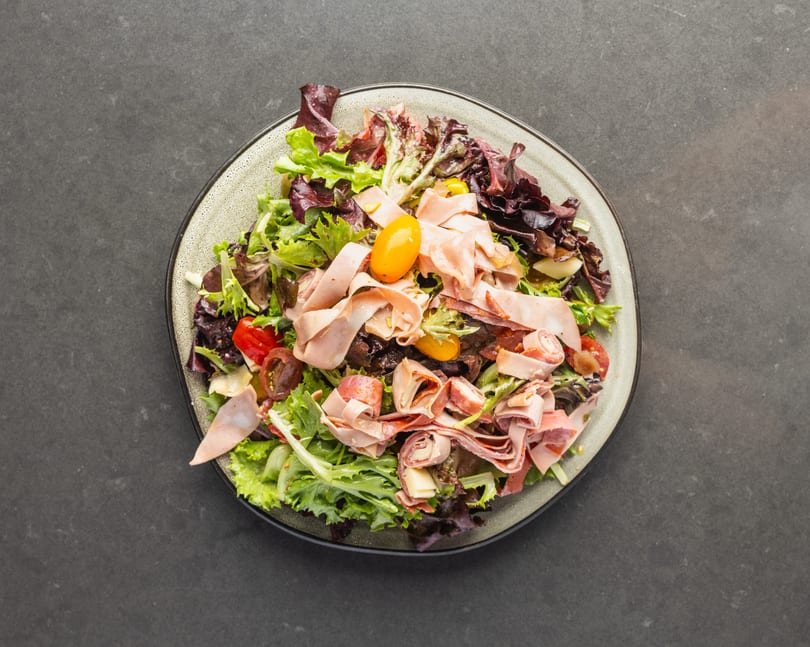 Overhead view of a salad with mixed greens, deli meats, cherry tomatoes, and cheese on a plate.