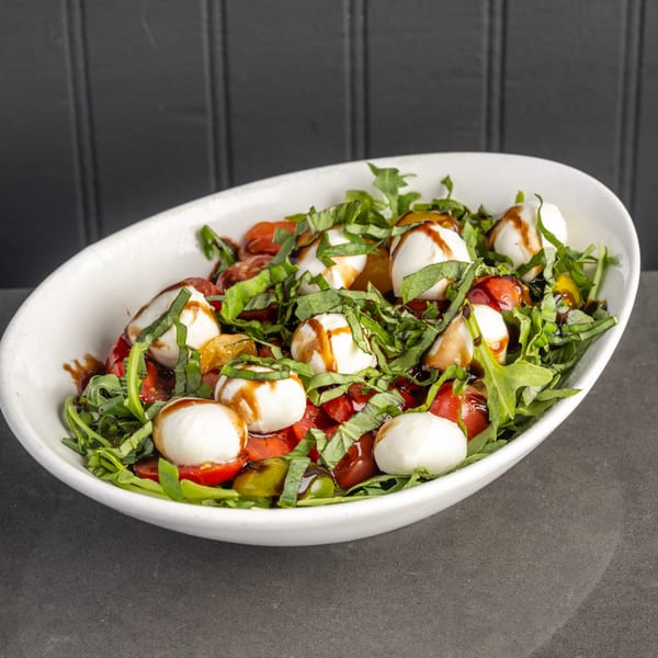 Caprese salad with fresh mozzarella, cherry tomatoes, basil, arugula, and balsamic glaze in a white bowl.