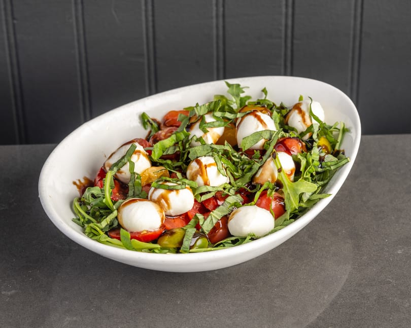 Caprese salad with fresh mozzarella, cherry tomatoes, basil, arugula, and balsamic glaze in a white bowl.